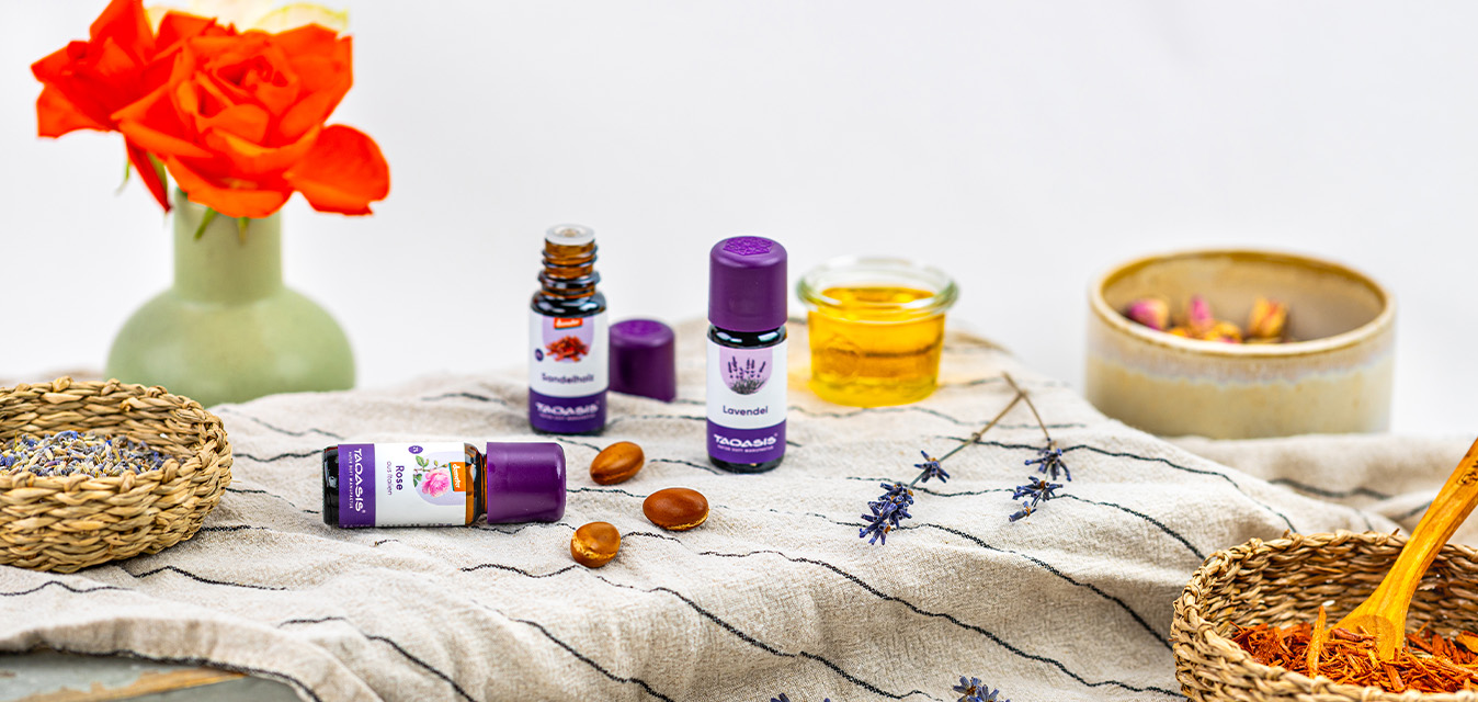 Ätherische Öle von TAOASIS mit Lavendel, Rose und Sandelholz, dekoriert mit Blüten, Pflanzenöl und natürlichen Zutaten auf hellem Leinentuch.