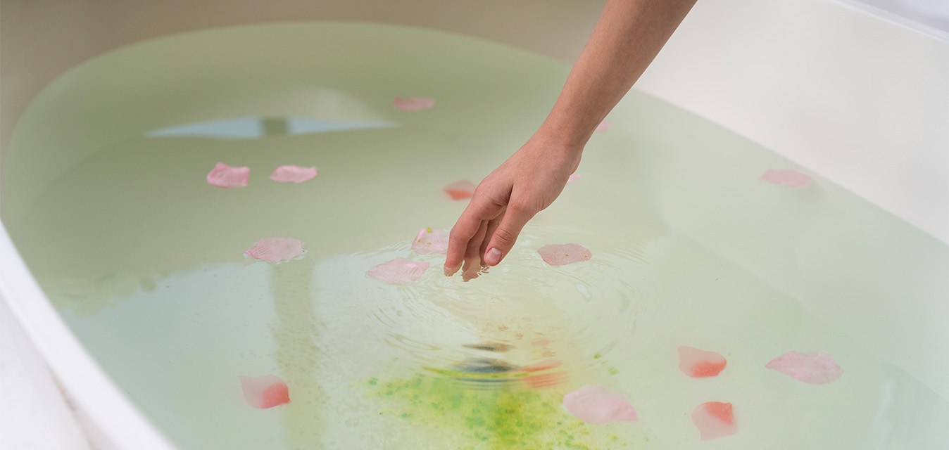 Hand berührt das Wasser eines entspannenden Vollbads mit schwimmenden Rosenblüten.