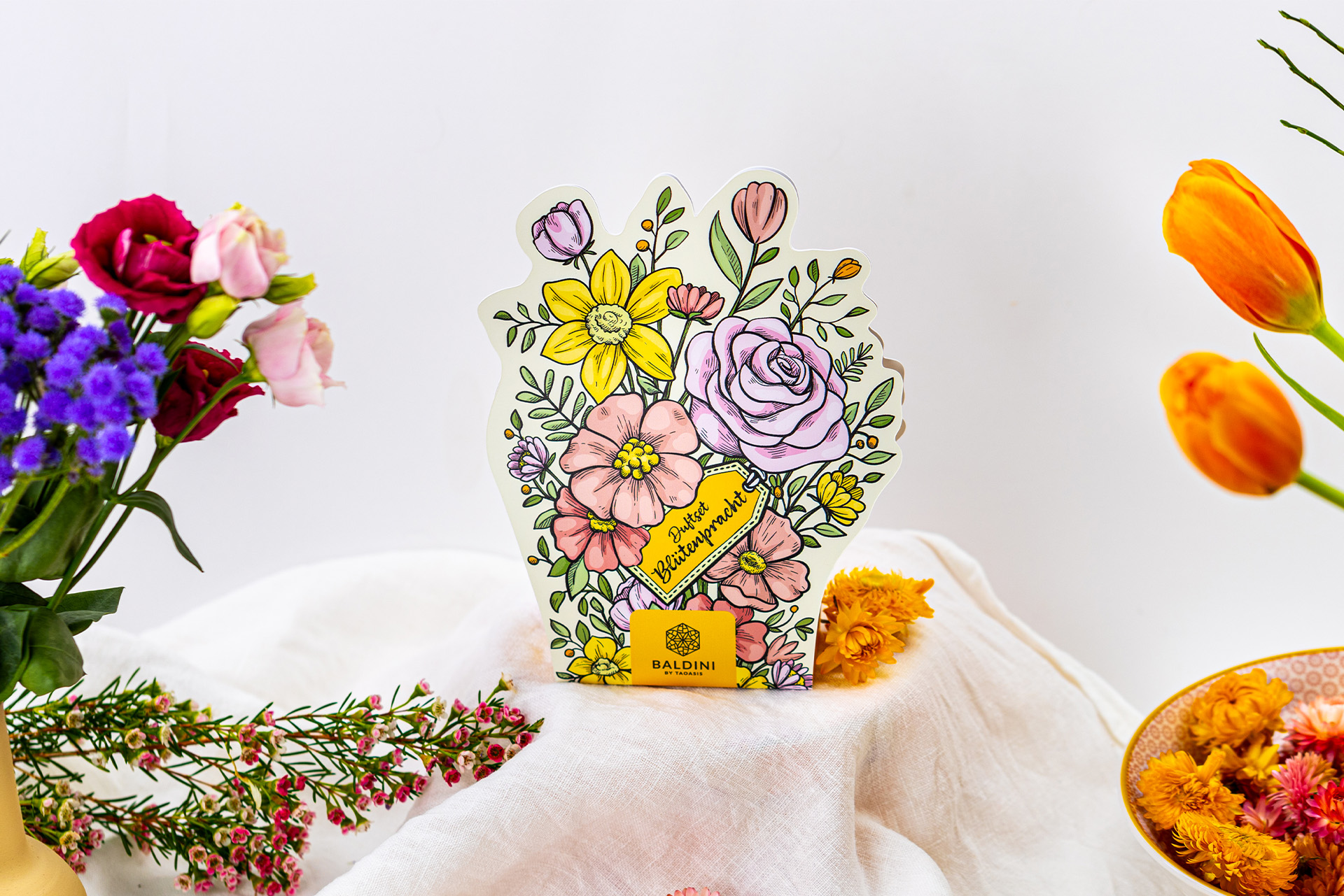 Das Baldini Duftset „Blütenpracht“ steht auf einem hellen Tuch und ist von bunten Blumen sowie getrockneten Blüten dekorativ umgeben.