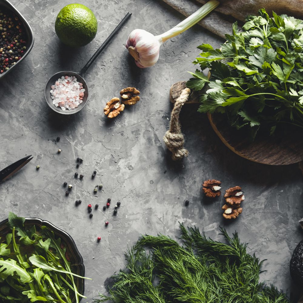 Frische Kräuter und Zutaten wie Petersilie, Dill, Rucola, Knoblauch, Limette, Walnüsse, Pfefferkörner und rosa Salz auf einer rustikalen Steinoberfläche arrangiert.