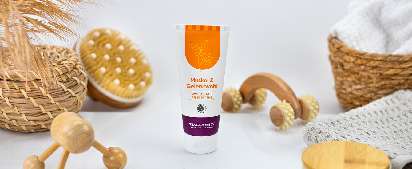 TAOASIS Muskel & Gelenkwohl Pflegecreme, umgeben von Massagehilfen, Handtüchern und natürlichen Wohnaccessoires.