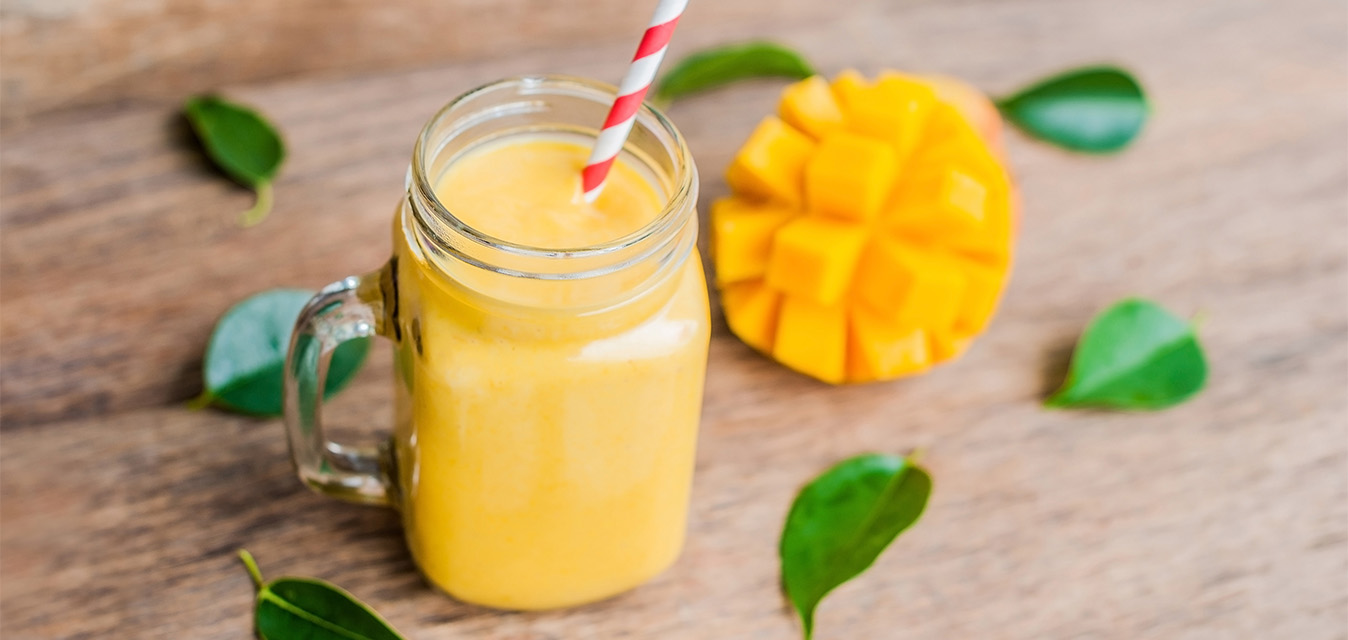 Mango-Smoothie im Glas mit Strohhalm, daneben eine aufgeschnittene Mango und grüne Blätter auf einem Holztisch.