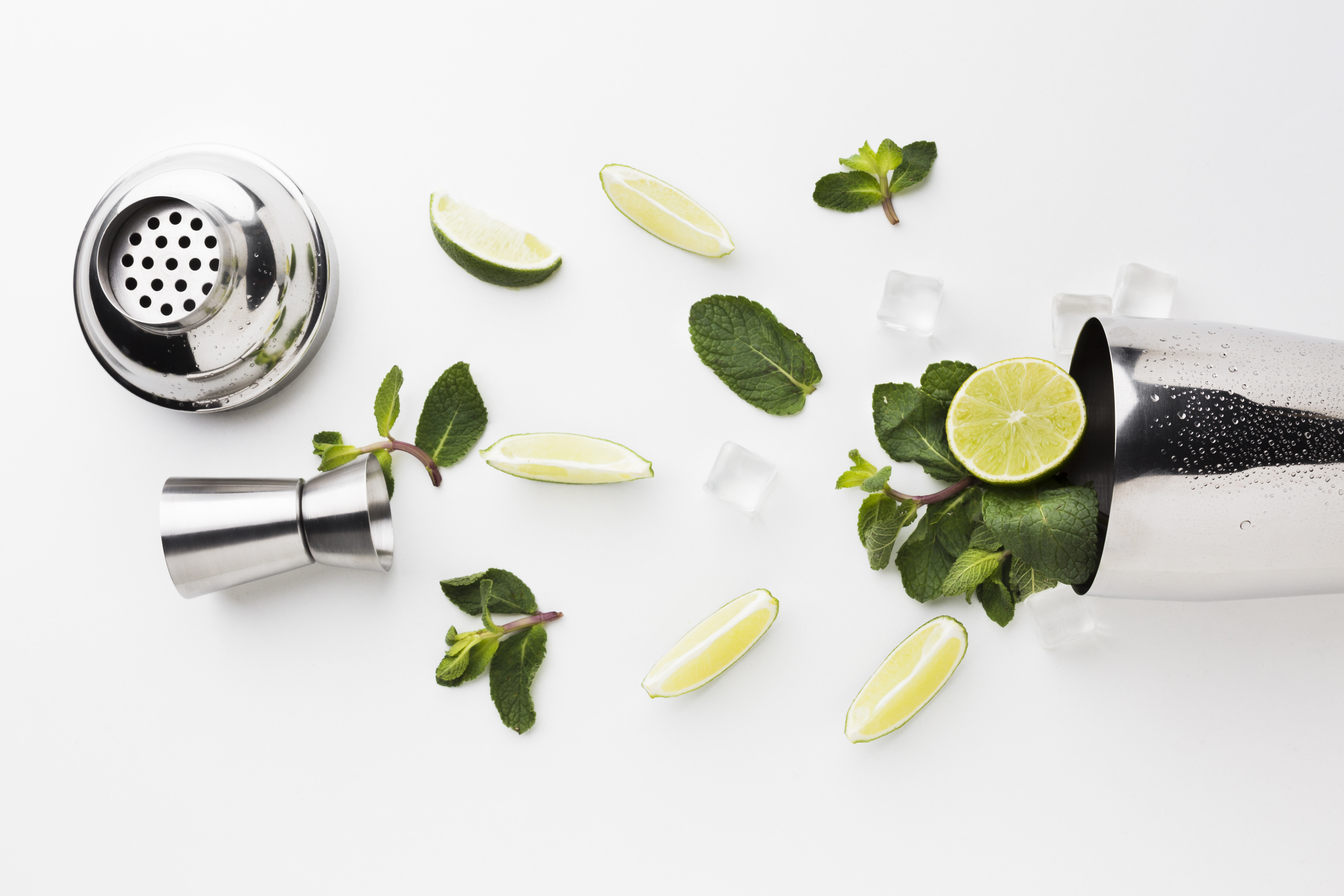 Cocktail-Zutaten flach arrangiert mit Shaker, Limettenscheiben, Minze und Eiswürfeln auf hellem Hintergrund.
