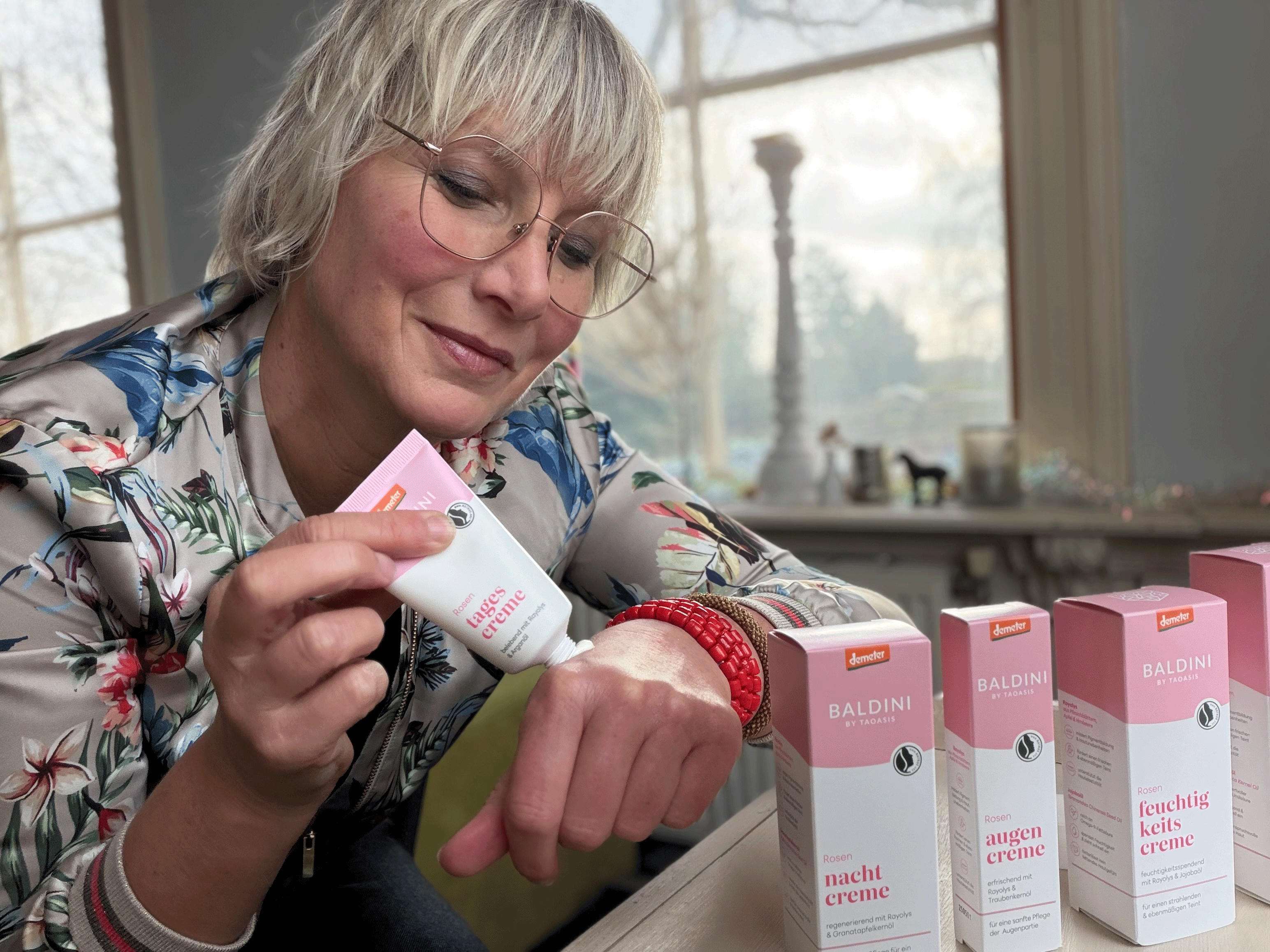 Frau trägt Rosencreme auf ihr Gesicht auf; im Vordergrund stehen mehrere Pflegeprodukte der Baldini Rosen-Serie.