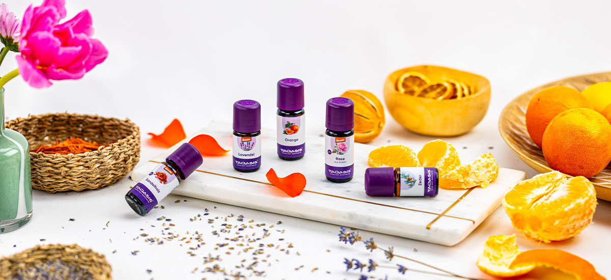 Verschiedene ätherische Öle von TAOASIS, darunter Lavendel, Orange, Rose und Zitrone, dekoriert mit frischen Orangen, Blüten und getrocknetem Lavendel.