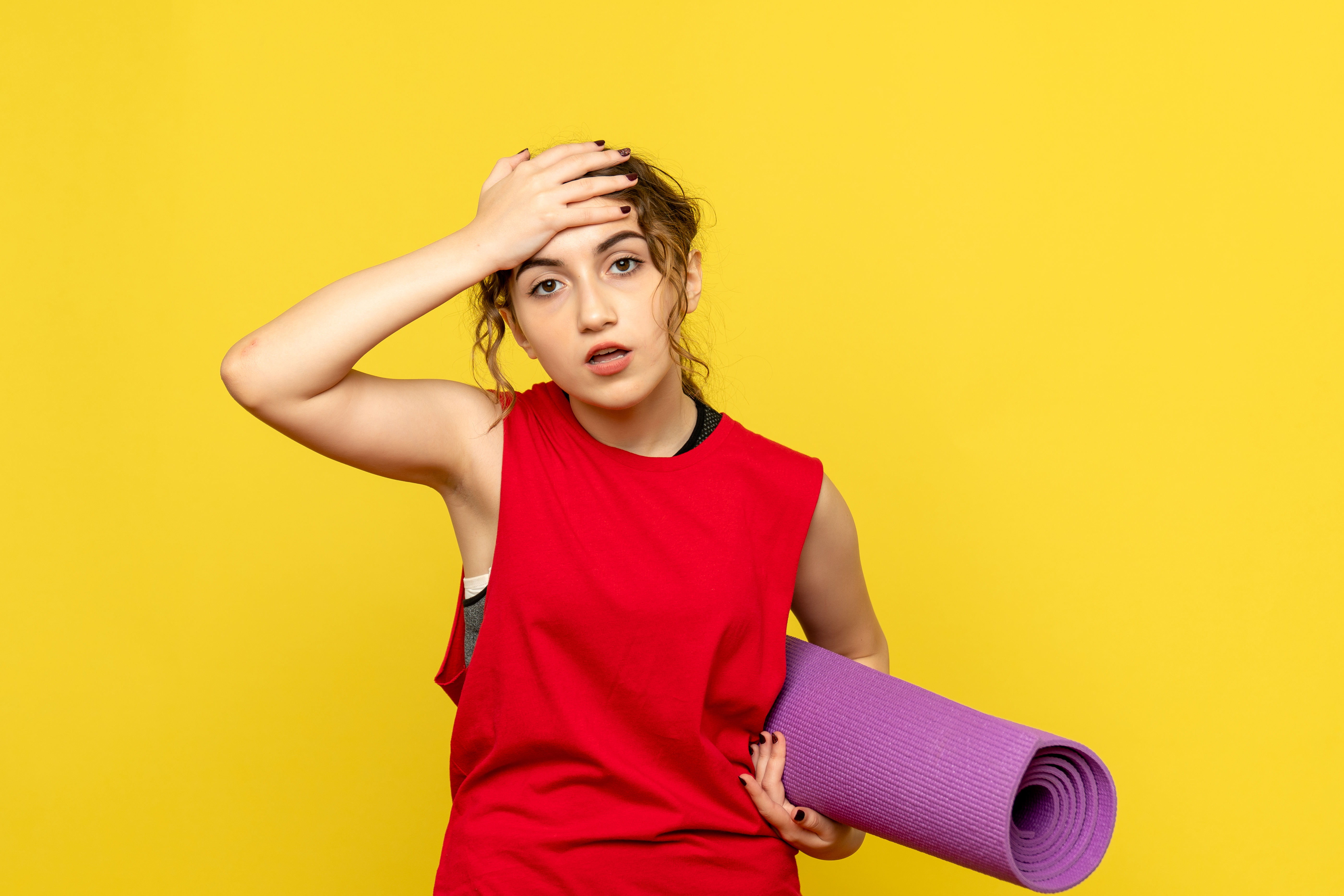 Junge Frau in rotem Sporttop mit Yogamatte vor gelbem Hintergrund wischt sich erschöpft über die Stirn.