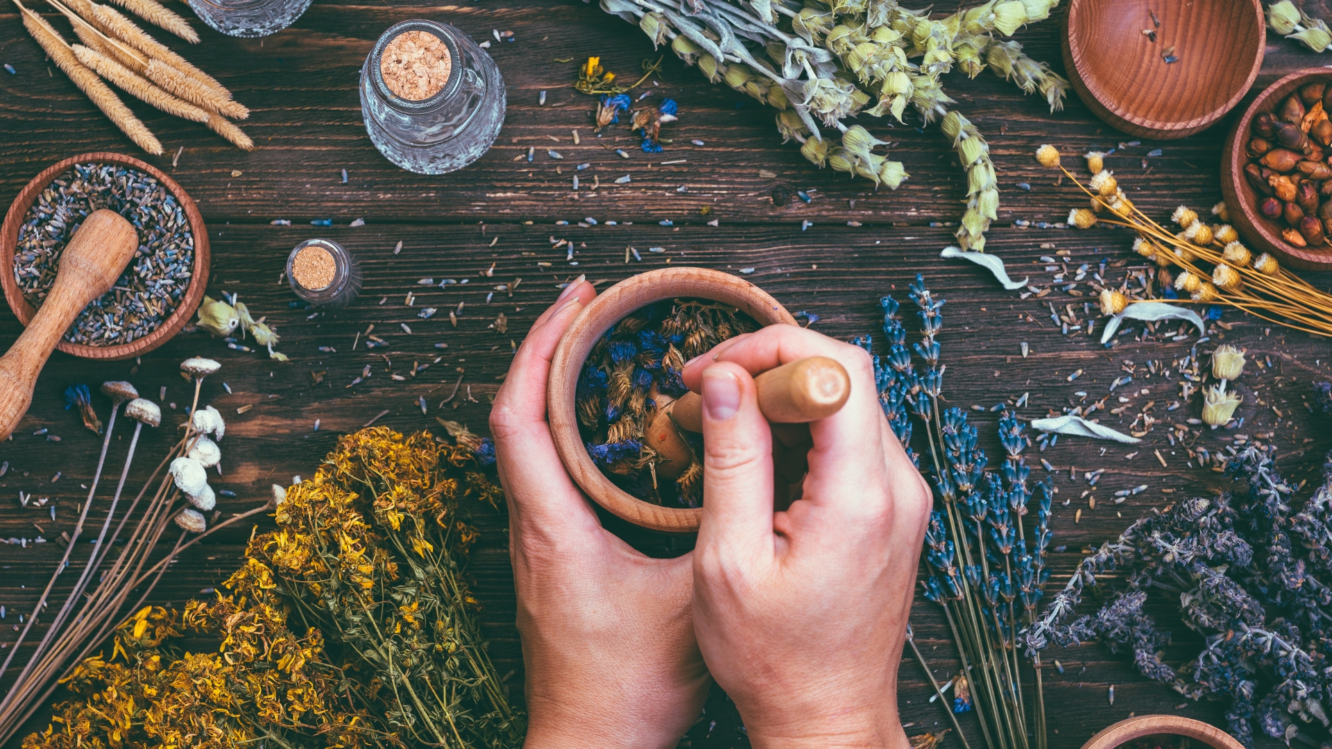 Ayurveda und Düfte Zwei Hände zerstoßen getrocknete Blüten in einer Holzschale mit Mörser, umgeben von verschiedenen Heilkräutern, Blüten, Samen und kleinen Glasfläschchen auf einem Holztisch – symbolisch für natürliche Heilkunst und ayurvedische Aromatherapie.