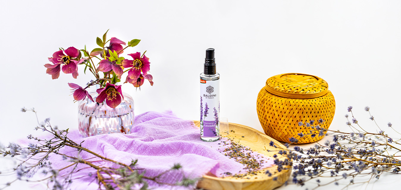 Baldini Lavendel Raumspray dekorativ mit violetten Blüten, getrocknetem Lavendel und lilafarbenem Tuch auf hellem Hintergrund.