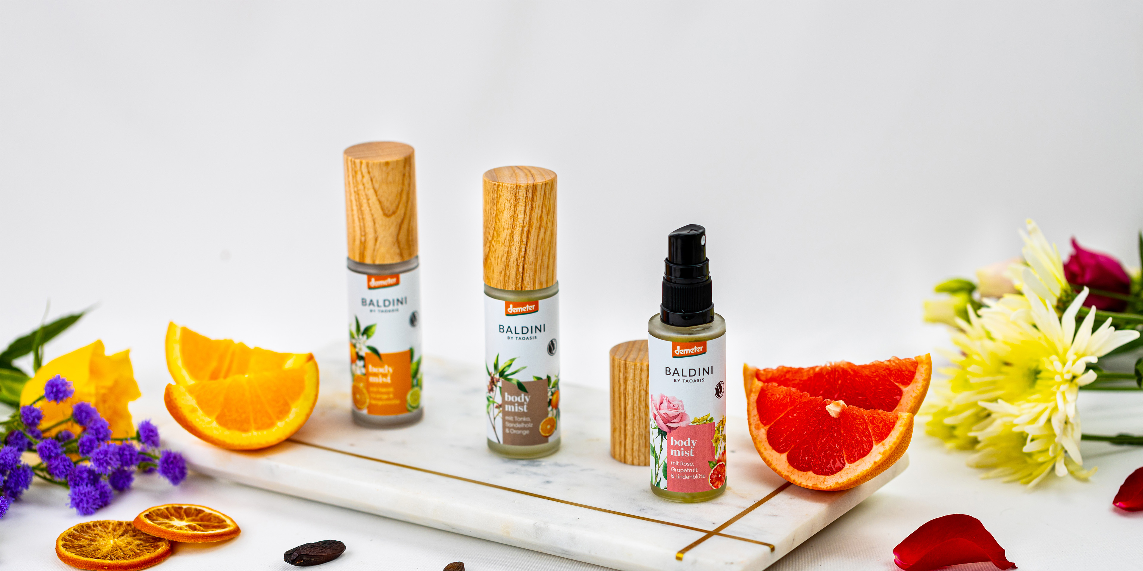 Baldini Body Mists in verschiedenen Duftvarianten, arrangiert mit frischen Zitrusfrüchten und bunten Blüten.