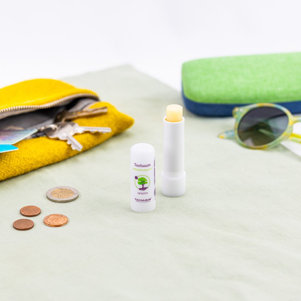 Teebaum Lippenpflegestift von TAOASIS neben Geldbörse, Münzen und Sonnenbrille.