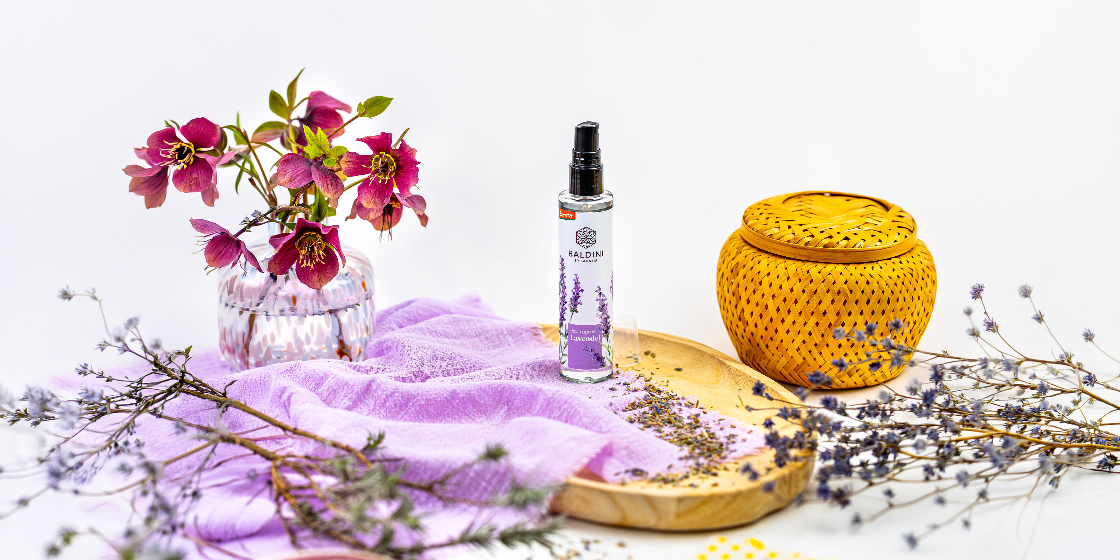 Baldini Lavendel-Raumspray auf einem Holztablett, dekoriert mit getrocknetem Lavendel, lilafarbenem Tuch, Blütenvase und geflochtenem Korb.
