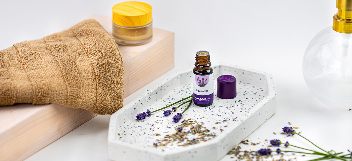 TAOASIS Lavendelöl auf einem hellen Tablett, dekoriert mit Lavendelblüten, getrocknetem Lavendel, Handtuch und Pflegeaccessoires.