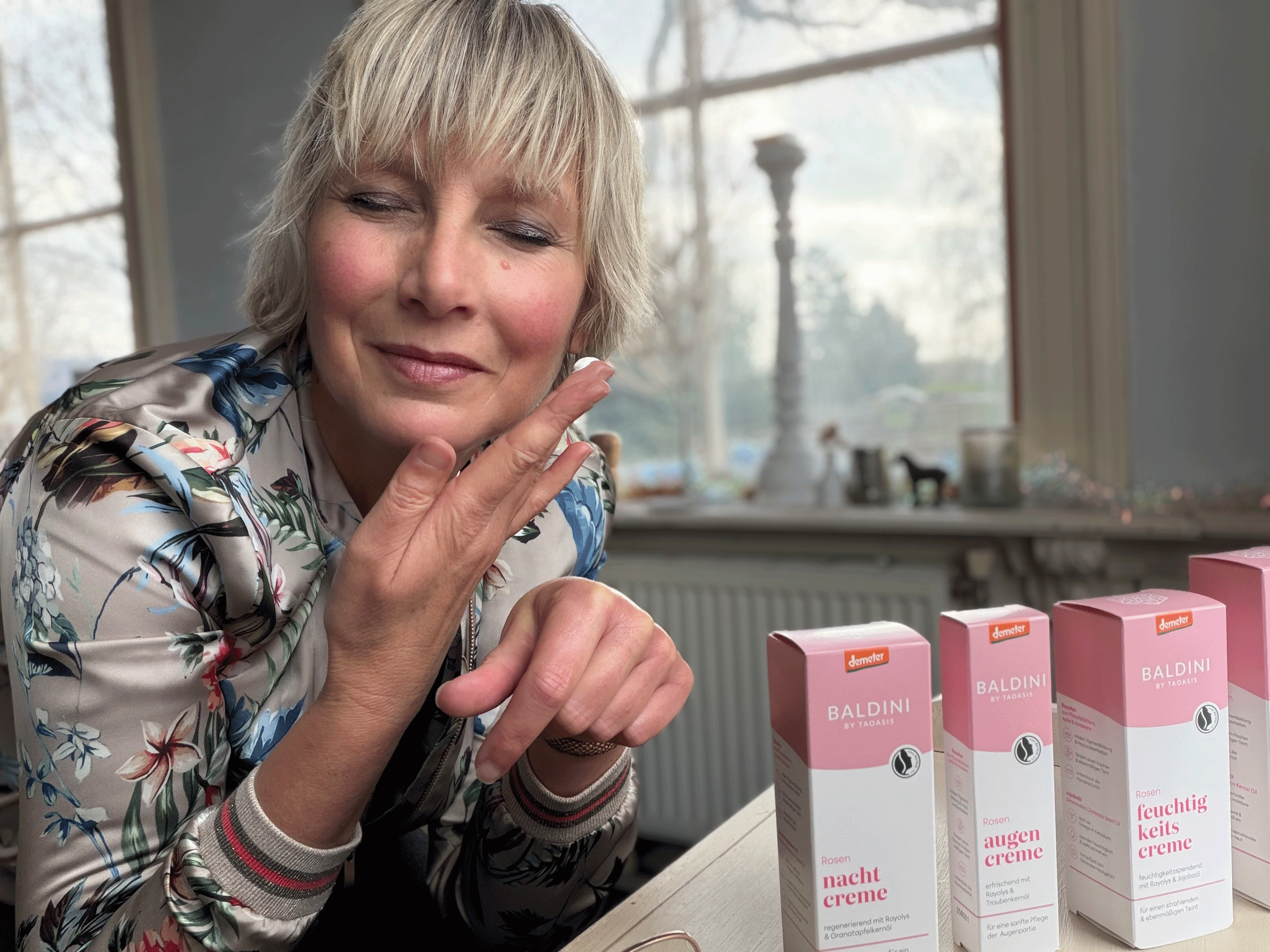 Frau trägt Rosen-Tagescreme auf ihren Handrücken auf; daneben stehen Pflegeprodukte der Baldini Rosen-Serie.
