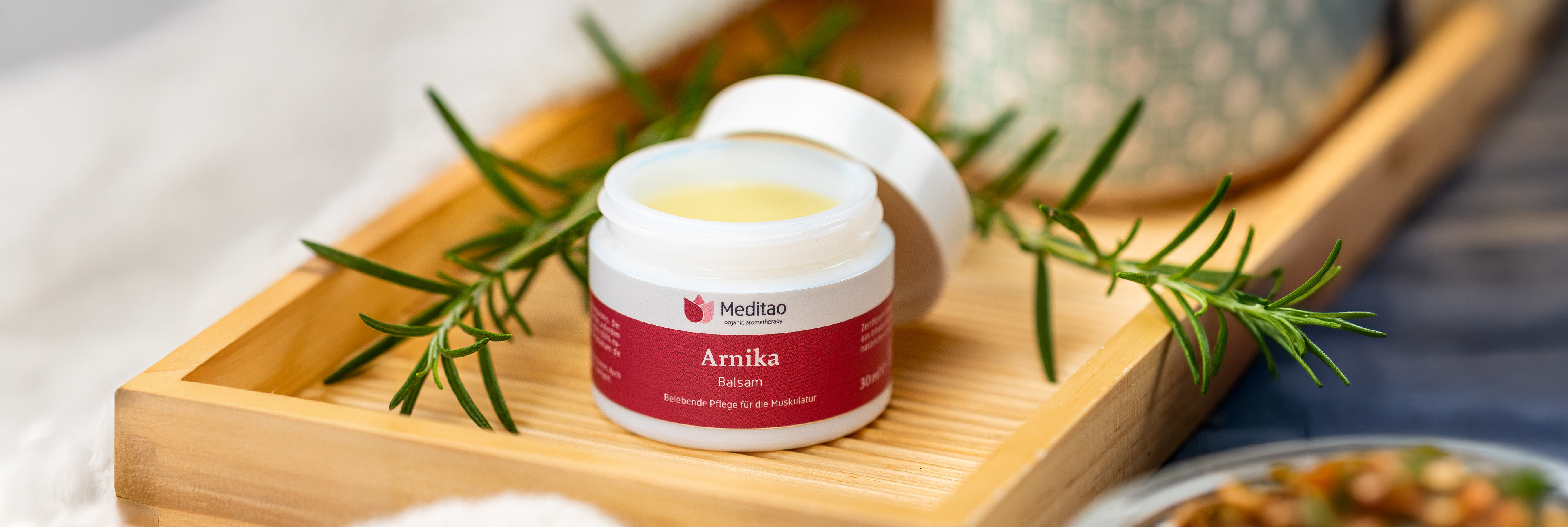 Meditao Arnika Balsam mit Rosmarinzweigen auf Holztablett