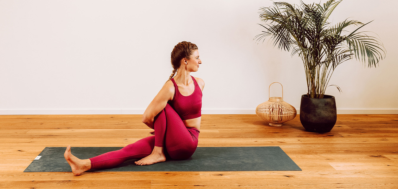 Frau in roter Sportkleidung bei einer Yoga-Drehhaltung auf einer Yogamatte in einem hellen Raum mit Holzboden und Zimmerpflanze.