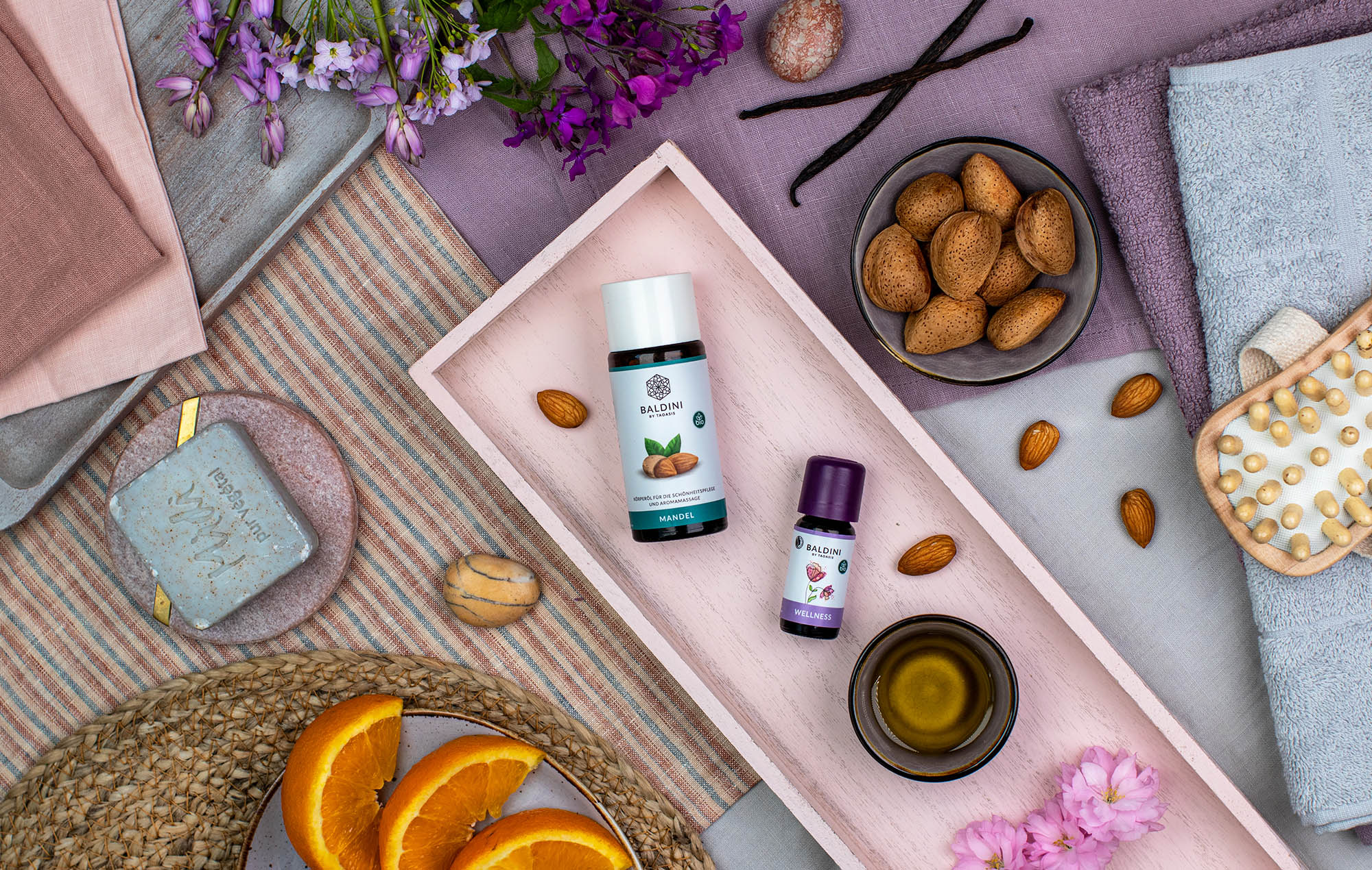 Wellnessöl mit Mandelbasis und ätherischem Duft, arrangiert mit Mandeln, Orangen, Blüten und Spa-Accessoires auf Tablett