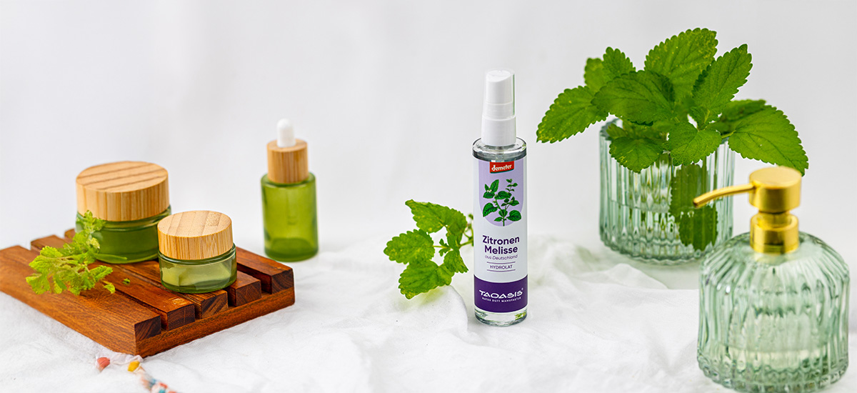 TAOASIS Zitronenmelisse Hydrolat, umgeben von frischer Zitronenmelisse, grünen Pflegefläschchen und natürlichen Badaccessoires.
