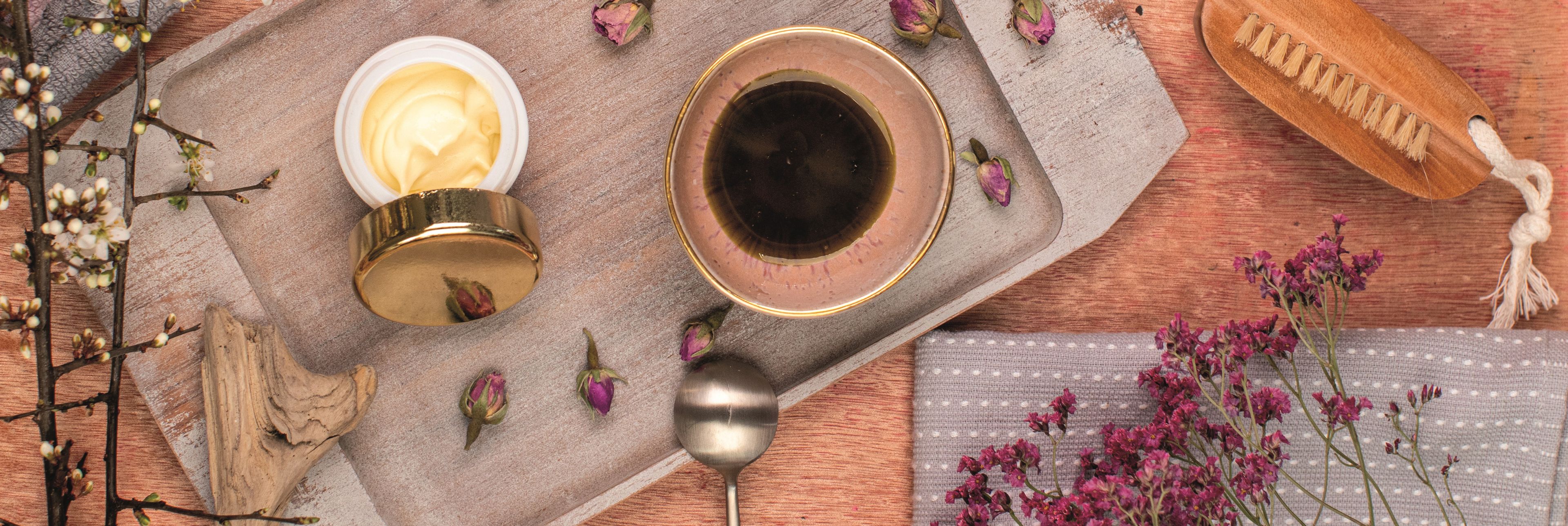 Das Bild zeigt eine liebevoll arrangierte Pflegeroutine mit Handcreme, Tee, Blüten und Naturmaterialien – warm, gemütlich und ideal für einen Moment achtsamer Selbstfürsorge.