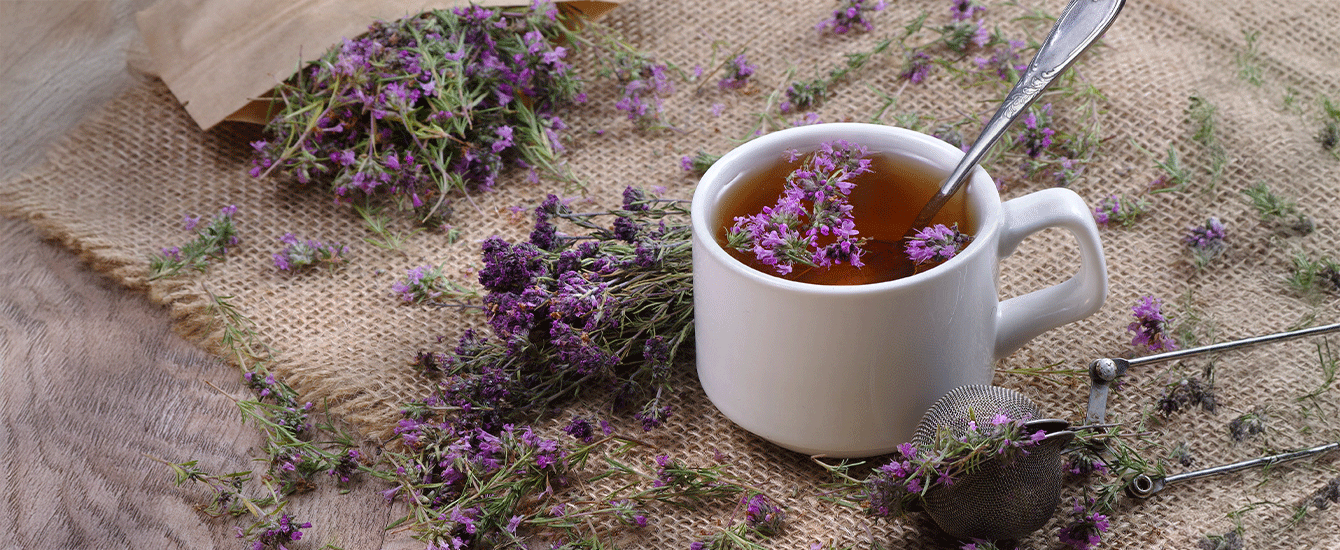 Tasse mit Kräutertee und blühendem Lavendel, umgeben von frischen Lavendelzweigen und einem Teesieb auf rustikalem Jutestoff.