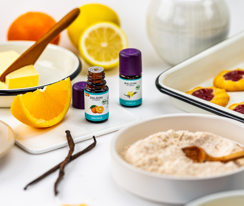 Bio Essenzen Orange und Vanille, im Hintergrund sind Kekse, man sieht Butter, Mehl und Zitrusfrüchte.