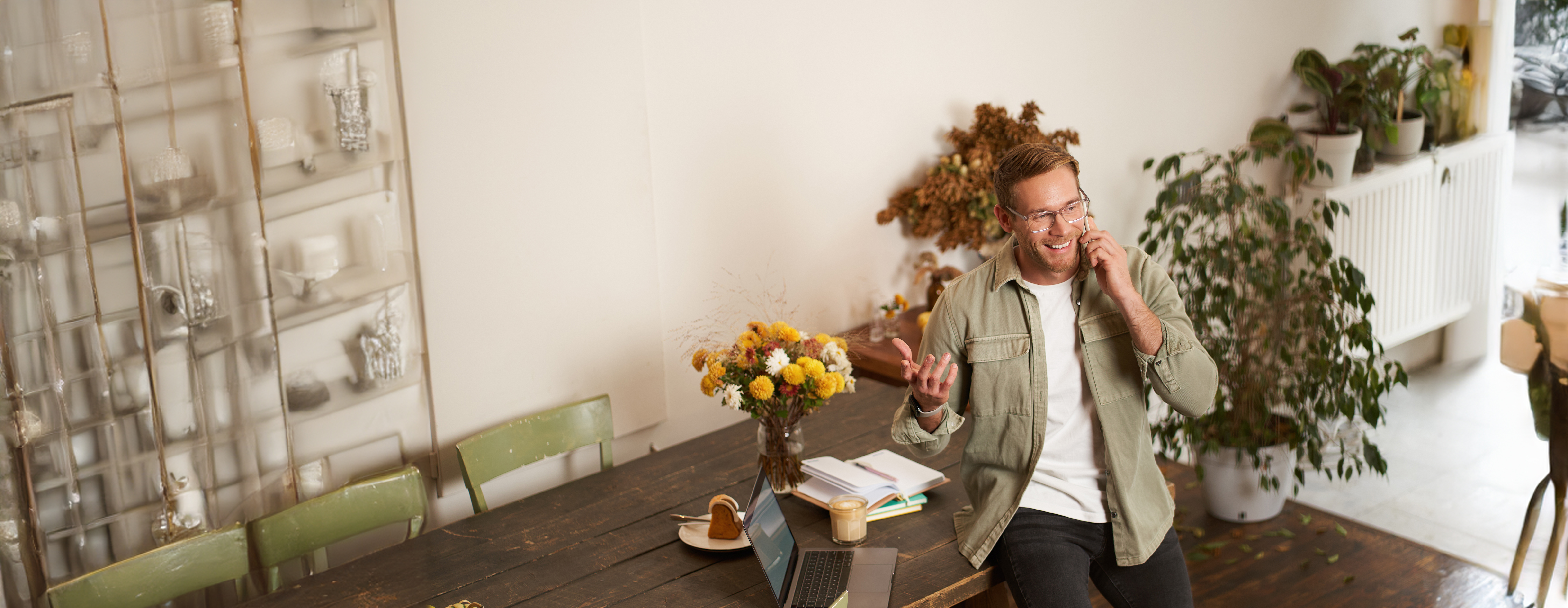 Mann sitzt an einem Holztisch mit Laptop und spricht lächelnd am Telefon in einem hellen, wohnlichen Raum mit Pflanzen.