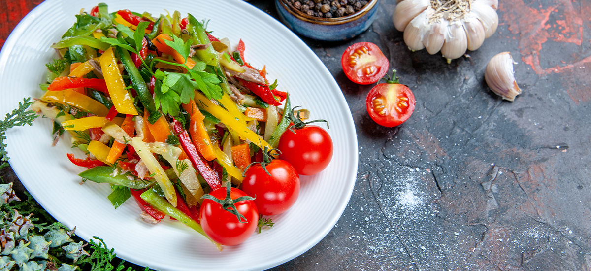 Bunter Gemüsesalat mit Paprika, Tomaten und frischen Kräutern auf einem weißen Teller, daneben Knoblauch, Pfeffer und Salz.