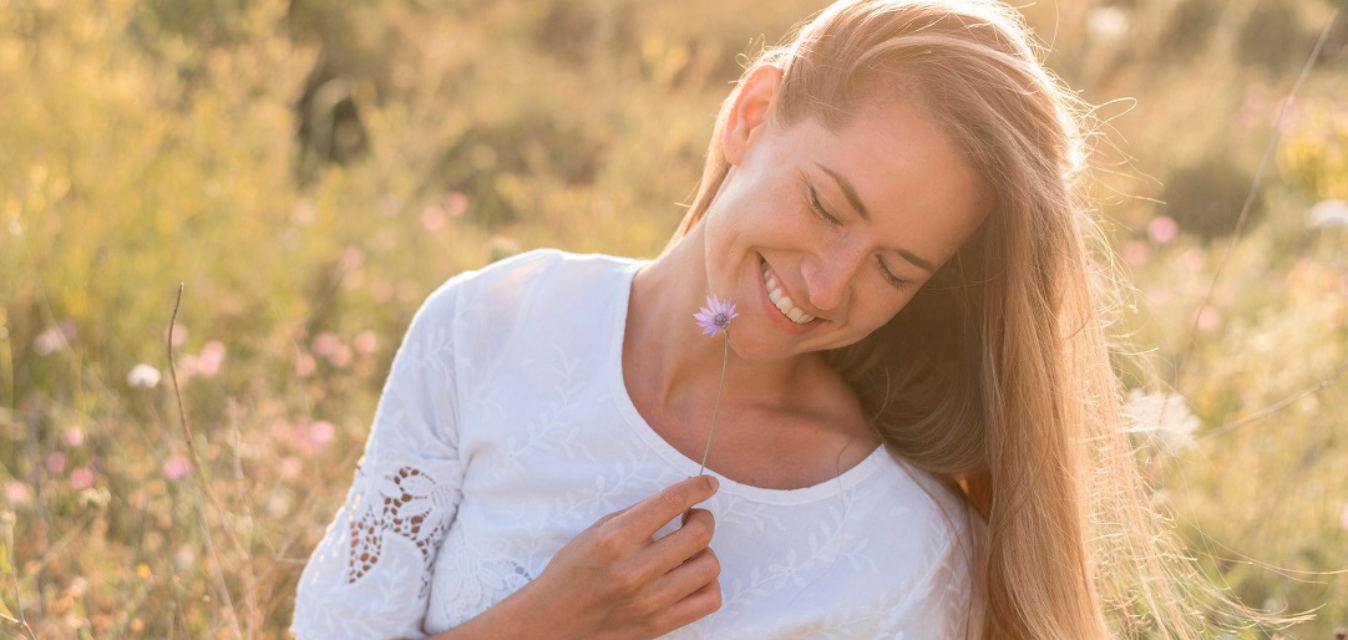 Frau hält eine kleine Blume und lächelt in einer sommerlichen Wiese.