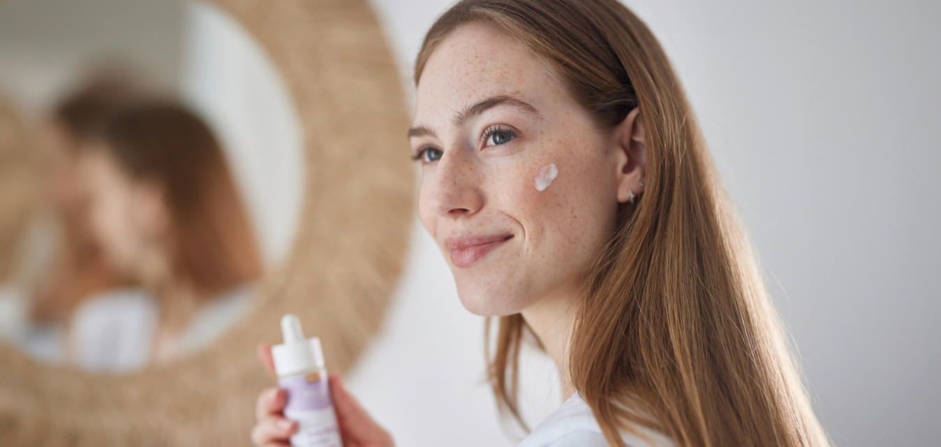 Frau mit Sommersprossen trägt Gesichtscreme auf und hält eine Naturkosmetikflasche