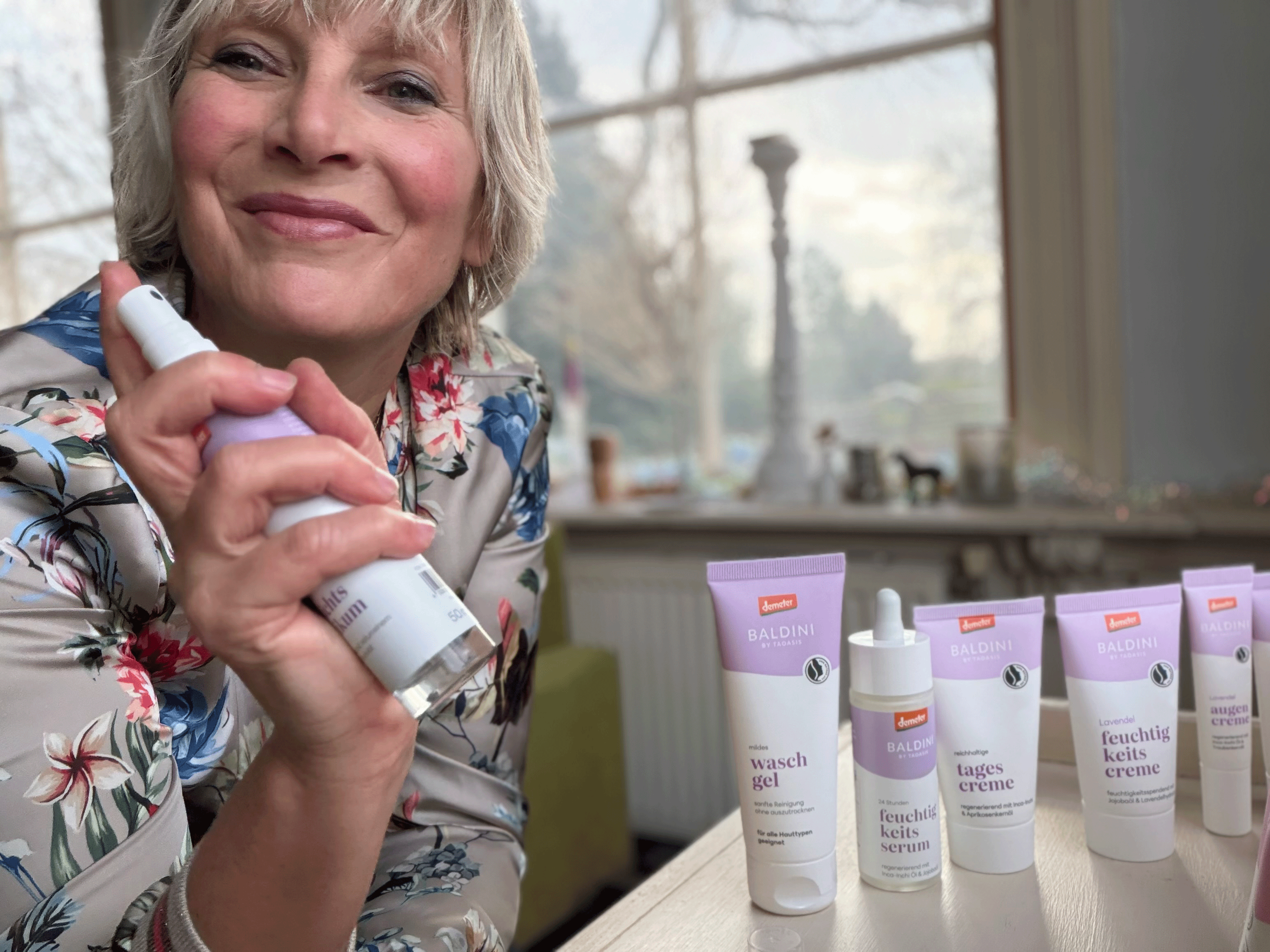 Frau präsentiert ein Baldini Lavendel-Raumspray; daneben stehen mehrere Produkte der Lavendel-Gesichtspflegeserie auf einem Tisch.