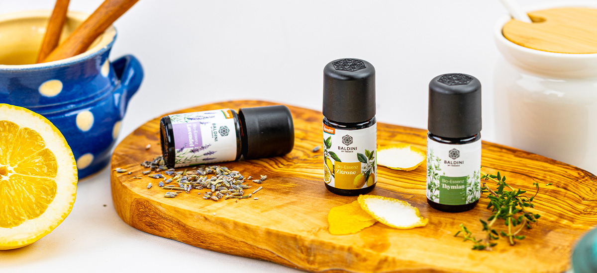 Drei Baldini Bio-Essenzen mit Lavendel, Zitrone und Thymian auf einem Holzbrett, umgeben von frischen Kräutern, Zitronenschale und Lavendelblüten.