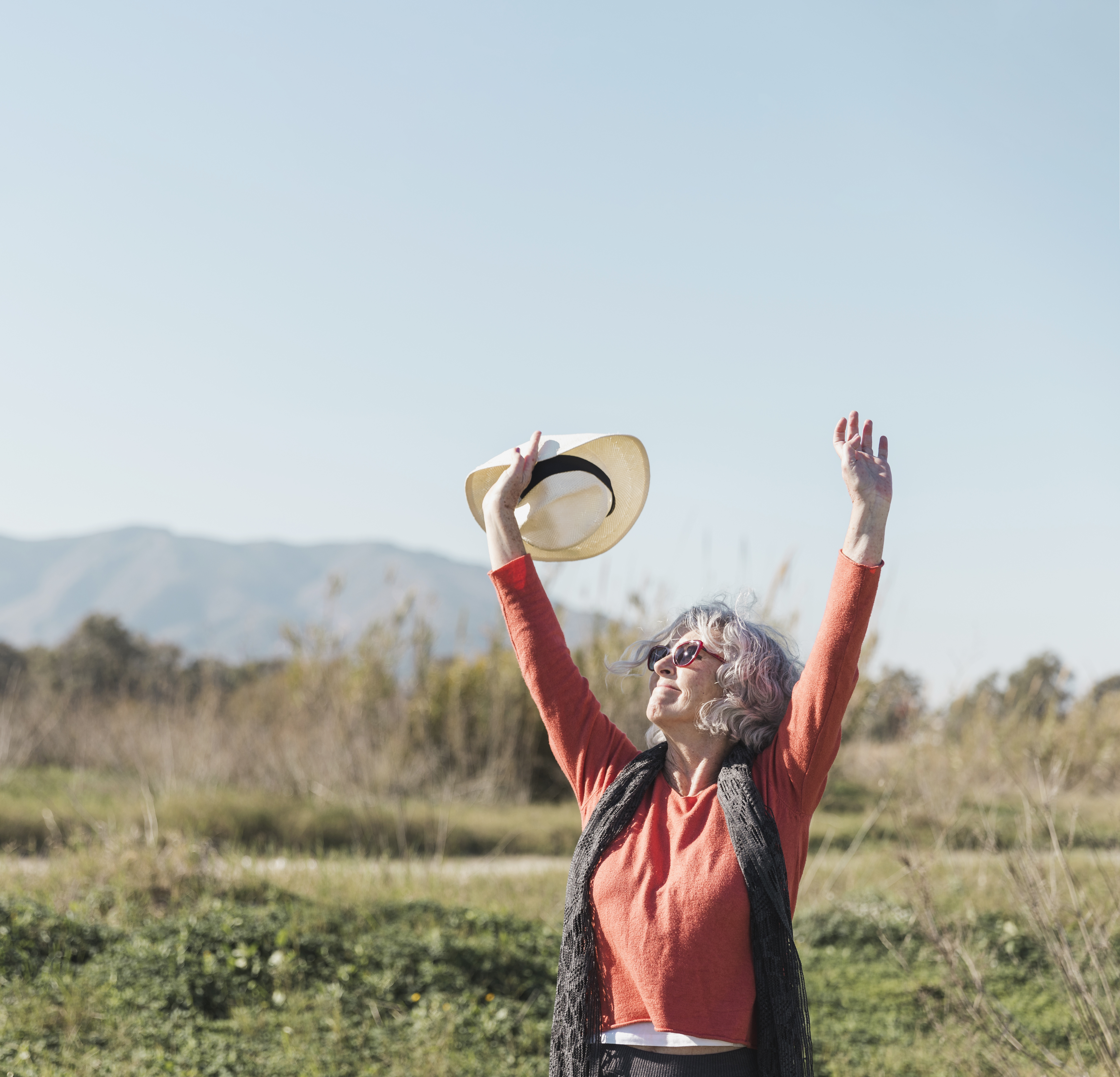 Frau mit Sonnenbrille hebt in der Natur die Arme und hält einen Hut in der Hand.