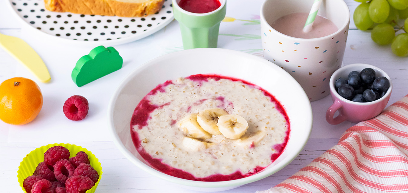 Warmes Porridge mit Banane und Beerensoße, umgeben von Himbeeren, Heidelbeeren, Trauben und einem Getränk auf einem hellen Frühstückstisch.